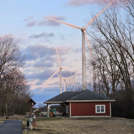 Red building with covered porch at trailhead of rail-to-trail park, with large wind turbines visible in the distance through bare trees.