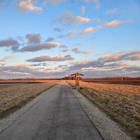 Eye-level view of a straight paved trail leading into the distance, with a tall, wooden bench on the right side offering a place to rest. The trail is flanked by dry, grassy fields under a blue sky with scattered clouds lit by the setting sun.
